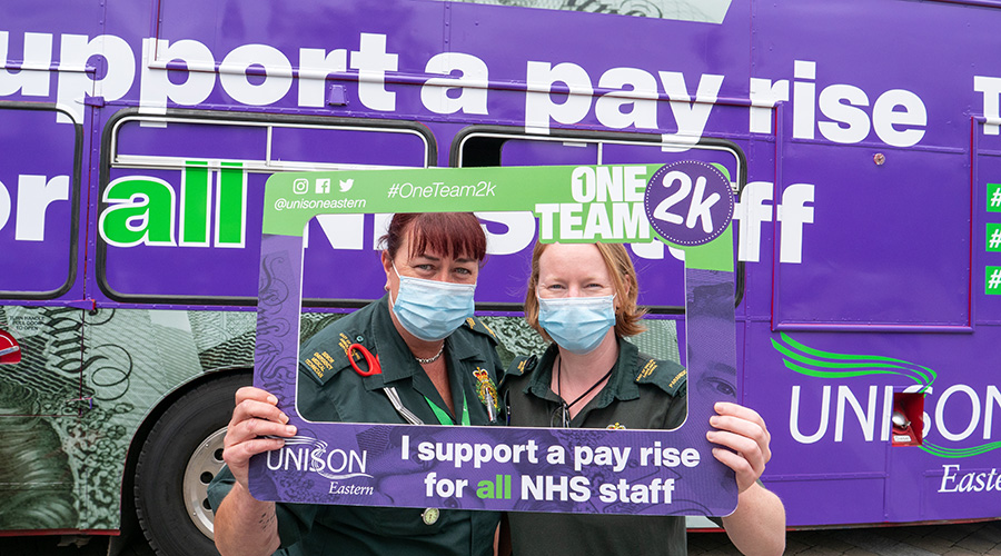 Two female ambulance workers hold a selfie frame supporting an NHS pay rise with a bus also supporting an NHS pay rise in the background