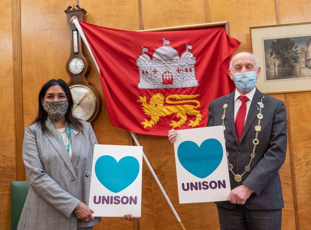 Debbie and the Lord Mayor hold Heart UNISON signs in front of the Norwich flag