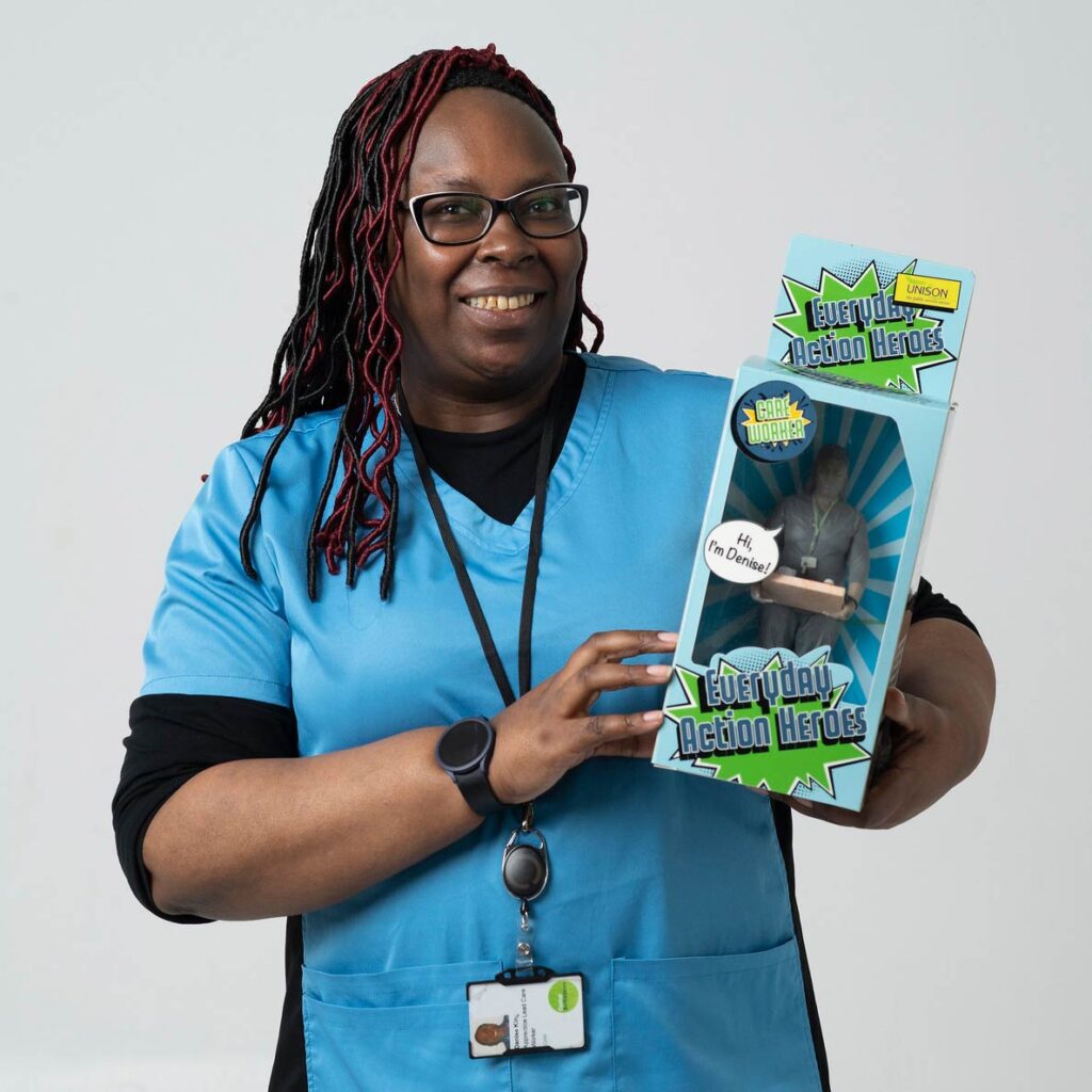 Dressed in her care uniform, Denise holds up a boxed action figure version of herself
