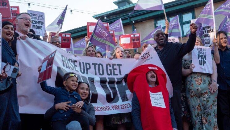 A man dressed as a postbox sits in front of a large group of people holding banners and UNISON flags. The message on the banner is 'Rise up to get pay up'
